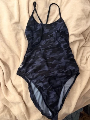 Mörkblå camo baddräkt från TYR - Snygg mörkblå baddräkt med camouflagemönster och tunna, korsade axelband. Baddräkten har en sportig design och öppen rygg, perfekt för simträning. Tillverkad i stretchigt material som sitter skönt på kroppen.