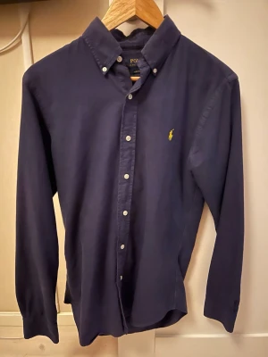 Skjorta från Polo Ralph Lauren - Klassisk mörkblå skjorta från Polo Ralph Lauren med button-down krage och den ikoniska gula loggan broderad på bröstet. Skjortan har långa ärmar, vita knappar och är tillverkad i mjuk bomull. Perfekt för en clean och stilren look.