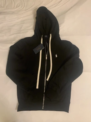 Svart hoodie från Polo Ralph Lauren - Svart hoodie från Polo Ralph Lauren med dragkedja framtill och vit broderad logga på bröstet. Hoodien har vita snören i huvan och ribbade muddar vid ärmar och nederkant. Tillverkad i mjuk bomullsblandning för en skön och avslappnad look. Mått: längd 60cm. Axelbredd 42cm.