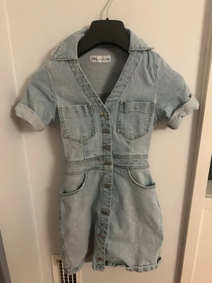 Ljusblå jeansklänning från Zara - Trendig ljusblå jeansbyxdress från Zara med korta ärmar och uppvik, knappar framtill och två bröstfickor. Byxdressen har även två sidofickor och en snygg krage. Perfekt för en avslappnad och cool look.knappt använd i storlek xs