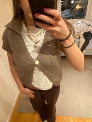 Stickad kofta - Kortärmad stickad kofta i brun nyans med stor krage och en knapp framtill. Koftan har en croppad passform och är gjord i ett mjukt, ull-liknande material. Perfekt att slänga över en t-shirt för en avslappnad men trendig look.