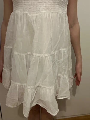 Vit kort sommarklänning med volanger - Söt vit kort klänning med tajt ribbning upptill och volangig kjol. Klänningen är ärmlös men har band där uppe och har en luftig passform, perfekt för varma dagar. Tillverkad i lätt bomullstyg som ger en avslappnad och fräsch känsla.