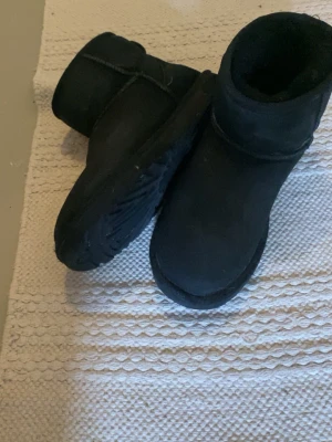 Svarta UGG boots i mocka - Klassiska svarta UGG boots i mjuk mocka med rund tå och kort skaft. Insidan är fodrad med fluffigt fårskinn för extra värme och komfort. Yttersulan är rejäl och greppvänlig, perfekt för kyliga dagar. Enkla att dra på och passar till många olika stilar.
