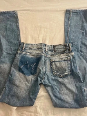Blå bootcut jeans med strassdetaljer - Säljer ett par ljusblå bootcut jeans från Mc Lem med snygga slitningar och strass/diamant detaljer på bakfickorna. Jeansen har låg midja, klassisk femficksmodell och är tillverkade i denim. Det är en snygg tvätt med markerade detaljer. Perfekta för dig som gillar en retro och trendig look.