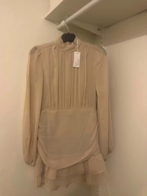 Beige volangklänning från Bubbleroom - Supersöt beige kortklänning från Bubbleroom med volangdetaljer och hög hals. Klänningen har långa ärmar, lätt puff vid axlarna och dragkedja i ryggen. Tillverkad i ett tunt, skirt material som ger en romantisk känsla. Perfekt för dig som gillar en feminin och trendig stil.