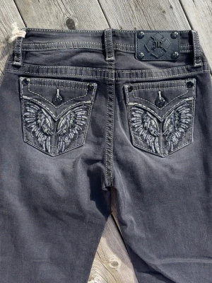 Gråa Miss Me jeans 26/32 - Snygga grå jeans från Miss Me med broderade fickor bak i form av vingar och dekorativa knappar. Jeansen har bootcut-modell och kontrastsömmar som ger en cool look. Materialet är denim. Innersöm ben 78cm, rak över midja 36cm.