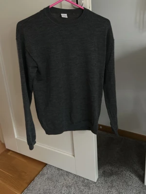 Mörkgrå stickad tröja - En enkel och stilren mörkgrå stickad tröja med rund halsringning och långa ärmar. Tröjan har ribbade muddar vid ärmslut och nederkant, perfekt för lager på lager. Passar till det mesta och är ett måste i garderoben.