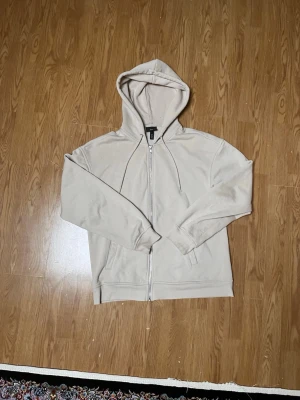 Beige hoodie med dragkedja från H&M - Snygg beige hoodie från H&M med dragkedja framtill och två fickor. Klassisk modell med huva och justerbara snören. Tillverkad i mjukt material som känns skönt mot huden. Perfekt för en avslappnad och stilren look.