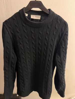 Svart kabelstickad tröja Selected - Snygg svart kabelstickad tröja från Selected med rund halsringning och ribbade muddar vid ärmslut och nederkant. Tröjan har ett klassiskt flätmönster och är perfekt för dig som gillar stilrena och tidlösa plagg. Materialet känns mjukt och bekvämt.