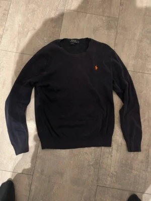 Mörkblå tröja från Polo Ralph Lauren - Snygg mörkblå tröja från Polo Ralph Lauren i slim fit-modell, tillverkad av mjuk Pima bomull. Tröjan har rund halsringning och den klassiska broderade loggan i orange på bröstet. Perfekt för en stilren och enkel look.