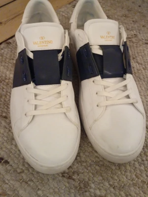 Valentino vita sneakers med blå detalj - Snygga vita sneakers från Valentino med bred mörkblå läderdetalj över snörningen och guldfärgad logga på plösen. Klassisk låg modell med vit sula och vita skosnören. Tillverkade i slätt läder för en clean och exklusiv look.