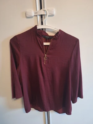 Vinröd blus med dragkedja  - Vinröd blus från Vero moda med trekvartsärm. Dragkedja framtill med rund metallring som detalj. Tillverkad i ett lätt, mjukt tyg som ger ett snyggt fall. Perfekt att styla med jeans eller kjol för en trendig look.
