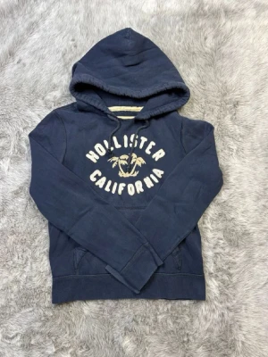Mörkblå hoodie från Hollister - Mörkblå hoodie från Hollister med tryck 'Hollister California' och palmer på bröstet. Klassisk huva och känguruficka framtill. Tillverkad i mjukt bomullsmaterial, perfekt för en avslappnad och cool stil.(storlek m)