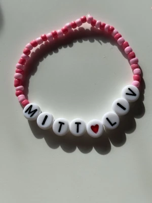 Pärlarmband med text MITT LIV - Sött armband med rosa och ljusrosa plastpärlor. De vita pärlorna har svarta bokstäver som bildar texten 'MITT LIV' och ett rött hjärta mellan orden. Perfekt för dig som gillar personliga accessoarer och färgglada detaljer.