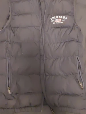 Mörkgrå dunväst från Polo Club - Snygg mörkgrå dunväst från Polo Club med broderad logga på bröstet. Västen har två dragkedjeförsedda fickor och quiltad design. Perfekt för lager-på-lager och håller dig varm under kyliga dagar. Klassisk och stilren look.