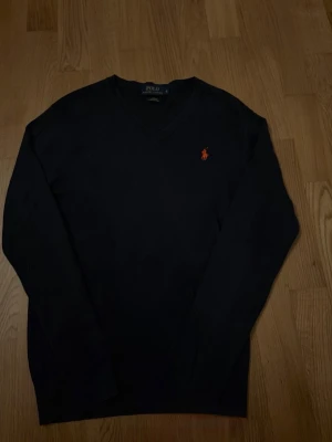 Mörkblå V-ringad tröja Polo Ralph Lauren - Mörkblå V-ringad tröja från Polo Ralph Lauren med den klassiska röda broderade loggan på bröstet. Tillverkad i mjuk bomull och har långa ärmar. Enkel och stilren design som funkar till det mesta.