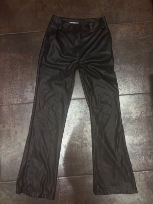 Svarta byxor från Madlady - Snygga svarta  byxor från Madlady med raka ben och hög midja. Byxorna har bälteshällor och knappstängning framtill. Materialet ger en cool läderlook som passar perfekt till streetwear eller en trendig outfit.