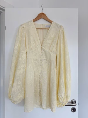 Ljusgul broderad klänning från Adoore -  Ljusgul kortklänning från Adoore med v-ringning, broderade blommor och klädda knappar framtill. Klänningen har långa puffärmar och markerad midja. Perfekt för sommarens alla tillfällen. Klänningen är aldrig använd och är därför i ett super fint skick💗