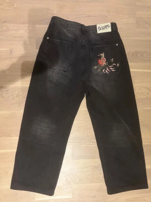 Derschutze jeans - Unika svarta jeans från Derschutze med rak passform och coolt tryck på bakfickan av en björn i röd tröja bland fiskar och löv. Jeansen har två bakfickor och är tillverkade i kraftig denim. Perfekta för dig som vill sticka ut med din stil. Storlek 28w men passar 30w, faller bra över skorna på mig som är 175