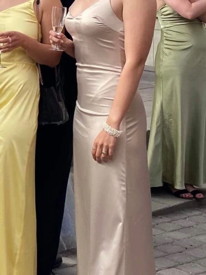 Champagnefärgad långklänning i satin - Elegant långklänning i champagnefärgad satin med smala axelband och figurnära passform. Klänningen har ett lätt glansigt material och djup skärning i ryggen. 