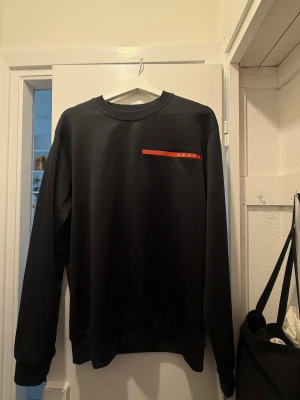Svart sweatshirt från Prada - Snygg svart sweatshirt från Prada med rund halsringning och en röd logodetalj på bröstet. Tröjan har långa ärmar och ribbade muddar vid ärmslut och nederkant. Perfekt för en stilren och modern look.