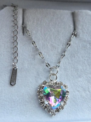 Halsband med regnbågshjärta och stenar - Snyggt halsband med ett hjärtformat hänge i regnbågsfärgad kristall, omgiven av gnistrande klara stenar. Kedjan är silverfärgad med dekorativa länkar. Perfekt för dig som gillar unika och glittriga smycken. Kedja i rostfritt stål 45 cm + 5 cm förlängningskedja.