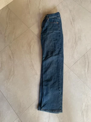 Blå raka jeans med fem fickor - Säljer ett par klassiska blå jeans med rak passform och fem fickor. Jeansen har normal midja, bälteshällor och stängs med dragkedja och knapp. Materialet är denim i bomull och färgen är en tidlös mellanblå nyans.