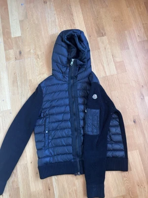 Svart pufferjacka från Moncler - Snygg svart pufferjacka från Moncler med stickade ärmar och vadderad kropp. Jackan har huva, dragkedja framtill och fickor med tryckknapp. Moncler-logga på ena ärmen och ribbade muddar för extra stilpoäng. Säljer ganska billigt för vill bli av med den, och ganska liten storlek passar ej mig längre 