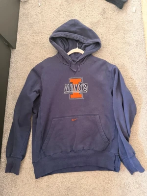 Blå Illinois Nike hoodie med huva Vintage - Snygg blå hoodie från Nike med stor orange Illinois-logga och text på bröstet. Klassisk känguruficka framtill, ribbade muddar och justerbar huva med dragsko. Perfekt för dig som gillar college-stil och sportiga plagg. Materialet känns mjukt och bekvämt.