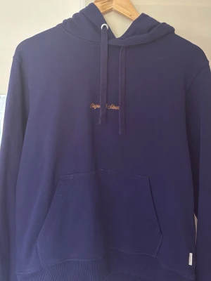 Mörkblå hoodie med broderad text från Jack and Jones - Mörkblå hoodie med huva och dragsnören, broderad text i gult på bröstet och stor känguruficka framtill. Tillverkad i mjukt bomullsmaterial och har ribbade muddar vid ärmslut och nederkant. Perfekt för en avslappnad och stilren look.