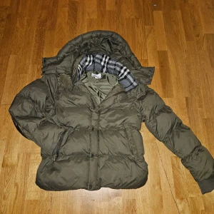 Burberry Pufferjacka - Säljer en olivgrön pufferjacka från Burberry med huva och klassiskt rutigt foder i svart, vitt och grått. Jackan har långa ärmar, ribbade muddar och stängs med både dragkedja och knappar. Perfekt för kalla dagar och riktigt snygg streetstil. Pris kan diskuteras lite. Finns ett väldogt litet hål på framsidan men inget som stör. Fraktar samma dag man köper.