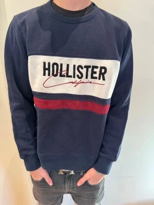 Blå Hollister Sweatshirr - Säljer en riktigt fet blå vit röd Hollister Hoodie i storlek M. Modellen på bilden är 180 cm lång och väger 65 kg.