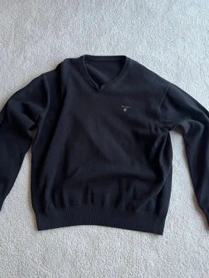 Svart stickad tröja från Gant - Klassisk svart stickad tröja från Gant med V-ringning och diskret logga broderad på bröstet. Tröjan har långa ärmar och ribbade muddar vid ärmslut och nederkant. Perfekt att matcha med skjorta eller t-shirt.