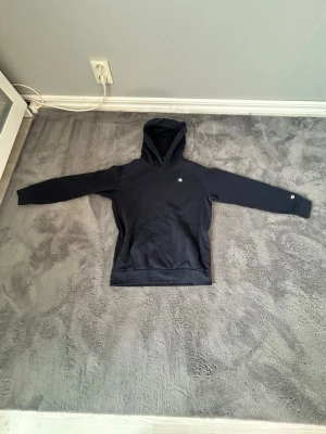 mörkblå hoodie från Champion - I storlek L i barn! Snygg mörkblå hoodie från Champion med klassisk logga på bröstet och ärmen. Hoodien har en stor huva och magficka framtill. Tillverkad i mjukt material som känns skönt mot huden. Perfekt för en avslappnad och sportig stil.