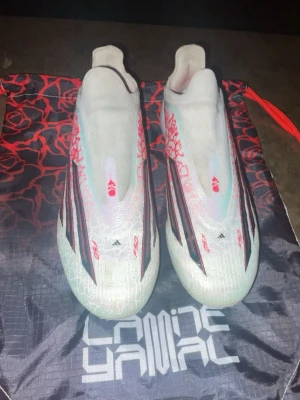 Ordinarie pris 3200 Adidas X fotbollsskor Lamine Edition - Snygga Adidas X fotbollsskor Elite i vitt med röda och svarta detaljer samt ros-mönster på sidorna. Skorna har låg profil, strömlinjeformad design och är tillverkade i syntetmaterial. Kommer med en svart och röd påse med Lamal Yamal-tryck. Ordinarie pris 3100kr 