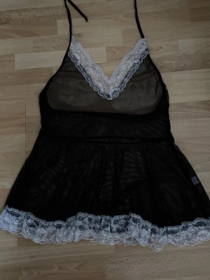 Ny. Svart transparent nattlinne med spets - Säljer ett svart nattlinne i transparent mesh med vita spetsdetaljer vid urringning och nederkant. Tunna axelband som knyts i nacken och v-ringad hals. Perfekt för dig som vill ha något både snyggt och bekvämt på kvällen.