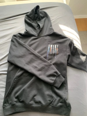 Svart hoodie från Amiri - Säljer en svart hoodie från Amiri med färgstänk-detaljer och logga på bröstet. Hoodien har klassisk känguruficka, ribbade muddar och en stor huva. Materialet känns mjukt och bekvämt, perfekt för en avslappnad streetwear-look.
