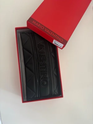 Svart plånbok från Mario Valentino - Snygg svart plånbok från Mario Valentino i quiltad design med tydlig logga på framsidan. Tillverkad i konstläder och levereras i original röd box. Perfekt för dig som gillar stilrena accessoarer med en lyxig touch.