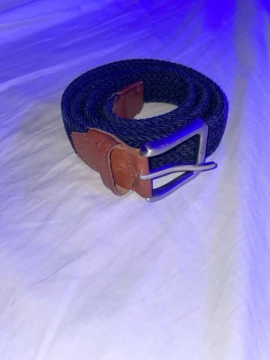 Mörkblått flätat bälte med läderdetaljer - Snyggt mörkblått flätat bälte med bruna läderdetaljer och silverfärgat spänne. Perfekt accessoar för att lyfta din outfit och funkar till både jeans och chinos. Enkel att justera och ger en stilren look.
