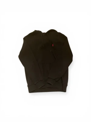 Svart hoodie från Ralph Lauren - Svart hoodie från Ralph Lauren med klassisk huva och dragsnören. På bröstet finns den ikoniska röda Polo-loggan broderad. Hoodien har en stor magficka och ribbade muddar vid ärmar och nederkant. Perfekt för en avslappnad och stilren look.