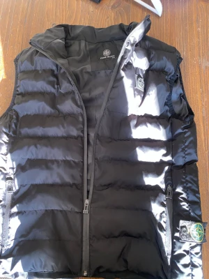 Svart dunväst från Stone Island - Snygg svart dunväst från Stone Island med ikonisk patch på vänster sida. Västen har två dragkedjeförsedda fickor, hög krage och quiltad design. Perfekt för lager-på-lager och streetwear-stil. Materialet är glansigt och lätt, vilket ger en modern look.