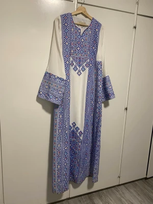 Vit långklänning med blått mönster - Unik vit långklänning med blått och rött broderat mönster. Klänningen har vida långärmar och en rak passform. Dekorativa detaljer längs ärmar, bröst och nederkant ger en traditionell och elegant look. Perfekt för dig som gillar statement-plagg. Det är en palestinsk klänning som har kommit till användning 1 kväll i 2 timmar.