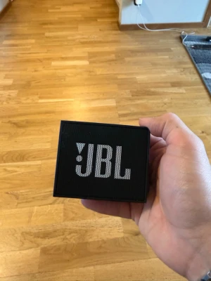 JBL svart bärbar högtalare - Smidig och kompakt svart bärbar högtalare från JBL. Högtalaren har en fyrkantig form och tydlig JBL-logga på framsidan. Perfekt för att ta med sig överallt och spela musik trådlöst. Tillverkad i slitstark plast med metallgaller.