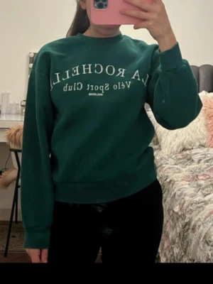 Grön sweatshirt LA ROCHELLA - Mörkgrön sweatshirt från LA ROCHELLA med vit text på bröstet. Tröjan har rund halsringning, långa ärmar och ribbade muddar vid ärmslut och nederkant. Perfekt för en avslappnad och sportig stil.