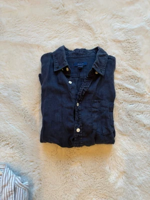 Mörkblå linneskjorta från Gant - Snygg mörkblå skjorta från Gant i mjukt linne. Klassisk krage, bröstficka och vita knappar framtill. Perfekt för en avslappnad men stilren look. Skjortan har lång ärm och en loose passform.