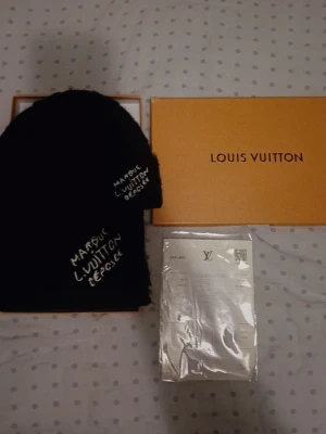 Louis Vuitton - Snygg svart mössa från Louis Vuitton med vit broderad text på sidan. Klassisk beanie-modell som ger en stilren och exklusiv känsla. Kommer med originalförpackning och tillbehör.