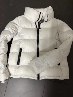 Colmar vit dunjacka - Vit dunjacka med puffig quiltad design och svarta dragkedjor. Hög krage och två sidofickor med dragkedja. Jackan ger en clean look. Perfekt för kallare dagar. 
