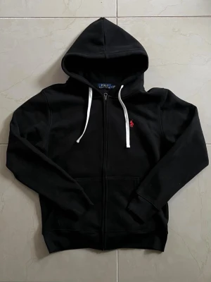 Svart hoodie från Polo Ralph Lauren - Säljer en svart hoodie från Polo Ralph Lauren med dragkedja, vit dragsko och röd logga på bröstet. Tröjan har känguruficka och mjukt material, perfekt för en avslappnad stil.