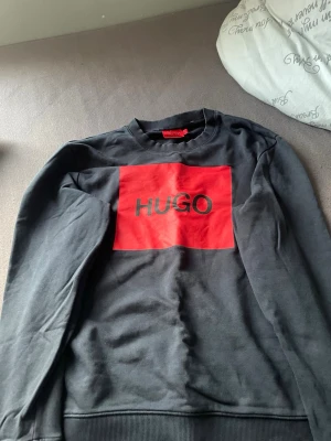 Svart sweatshirt från HUGO med röd logga - Svart sweatshirt från HUGO med stor röd ruta och svart HUGO-tryck på bröstet. Tröjan har rund halsringning, långa ärmar och ribbade muddar. Tillverkad i mjukt material som är skönt att bära och ger en clean streetwear-look.