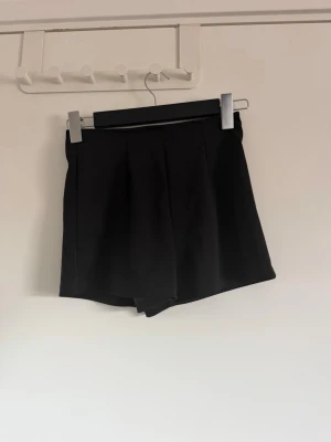 Svarta shorts med dragkedja bak - Stilren svart shorts med hög midja och dragkedja bak. Shortsen har en klassisk A-linjeform och är sydd i ett mjukt, lite stretchigt tyg som ger en snygg passform. Perfekt för dig som vill ha kjol-look men med shortskomfort.